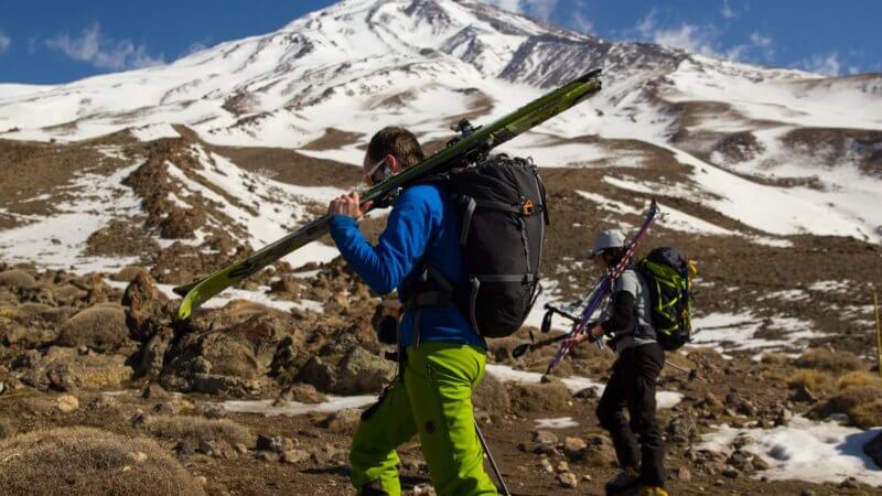 George and Shirin walk past the camera holding skis over one shoulder and trekking poles in the other. Photo of Mount Damavand behind.