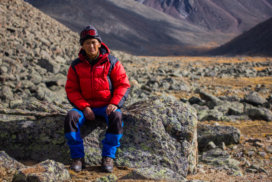 A Tuvan mountaineer wearing blue trousers and red jacket sat on a rock posing for the camera.