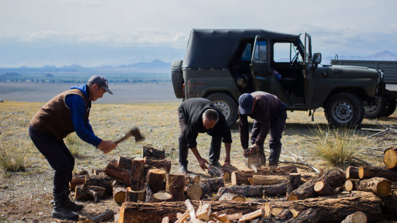 Three men next to a green Russian jeep chopping wooden logs.