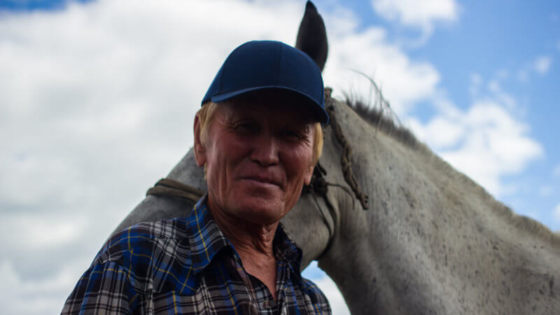 A man with blue cap and blue-white checked shirt smiles in front of a white horse.