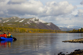 A man paddling a blue packraft descends the Yenisei River with snow-covered mountains behind.
