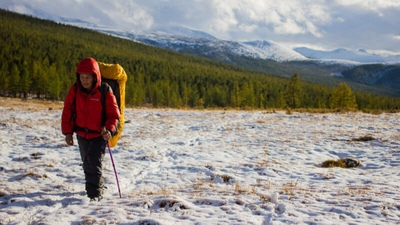 A hiker in red jacket and wearing a yellow bag walking through the snow.