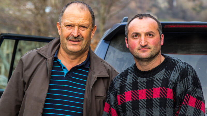 Two Albanian men pose for a photo in front of a car.