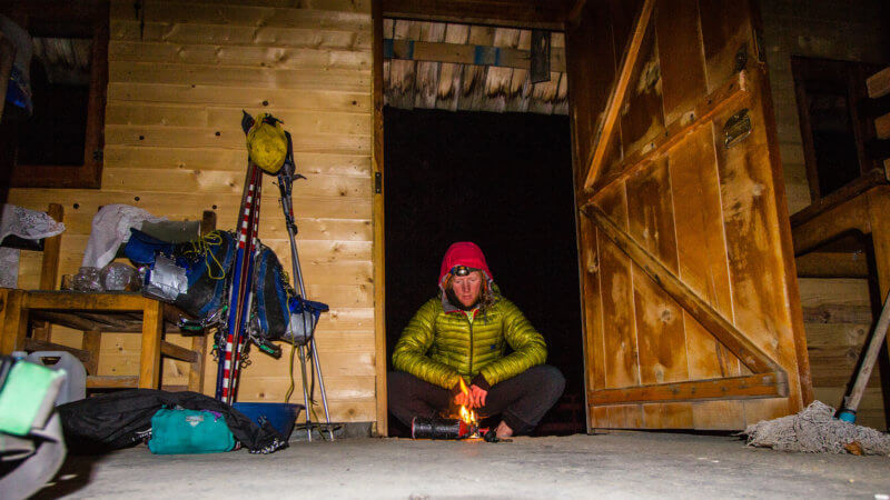 A camper with green jacket in mountain hut huddled around a petrol stove fire.