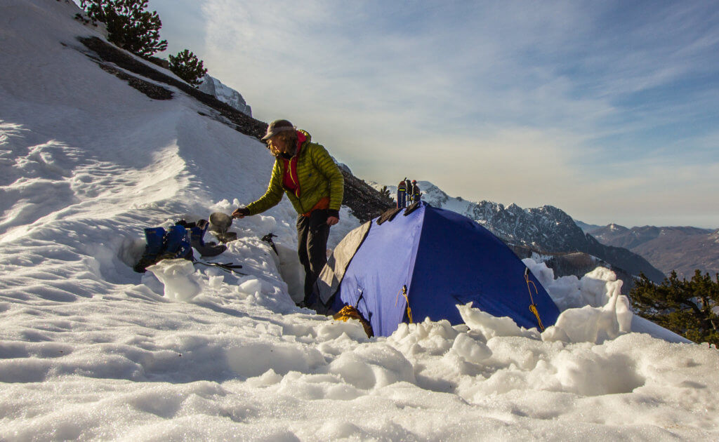 Skier camping atop snowy mountain pass with blue tent.