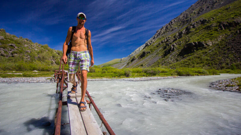 A shirtless mountain guide wearing sandals walks across a narrow footbridge spanning a glacial river.