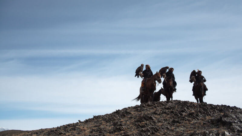 A far shot of three eagle hunters stood on top of a hillside with their horses.