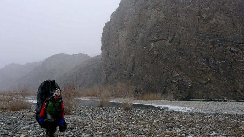 Jamie walking beside the Khovd River in winter with a packraft on his back.