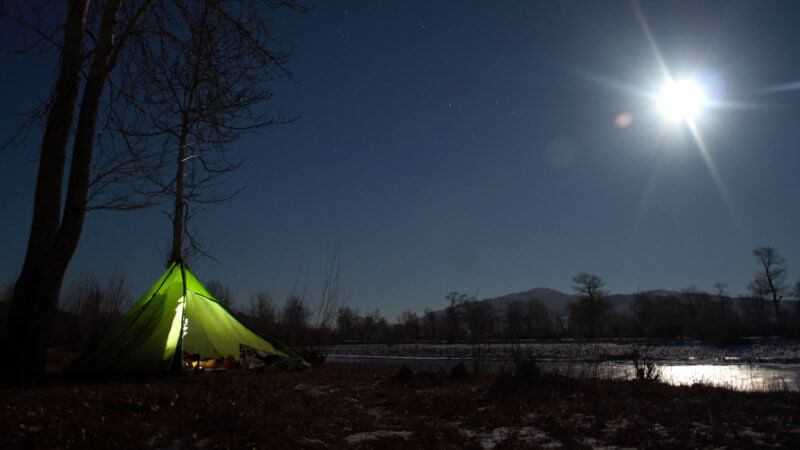 A night time shot of a green tent beside a frozen river in moonlight.