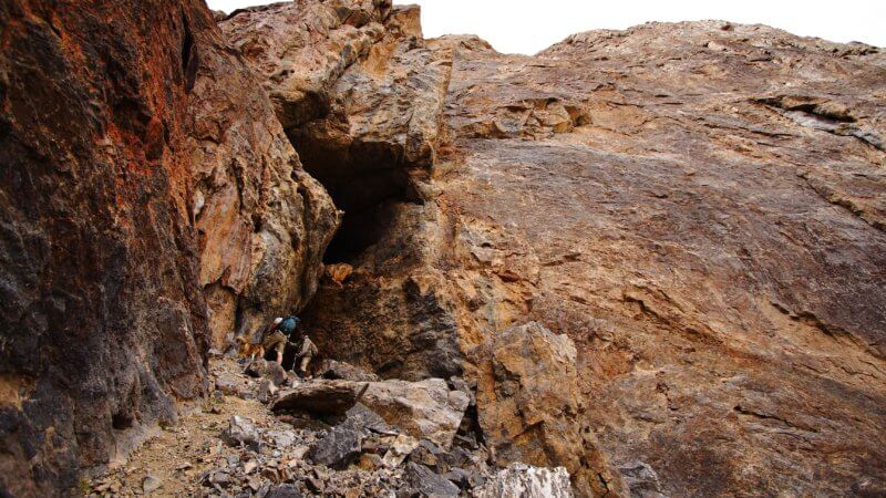 Two men and a dog scramble up to the entrance of a Pamir Mountain cave.