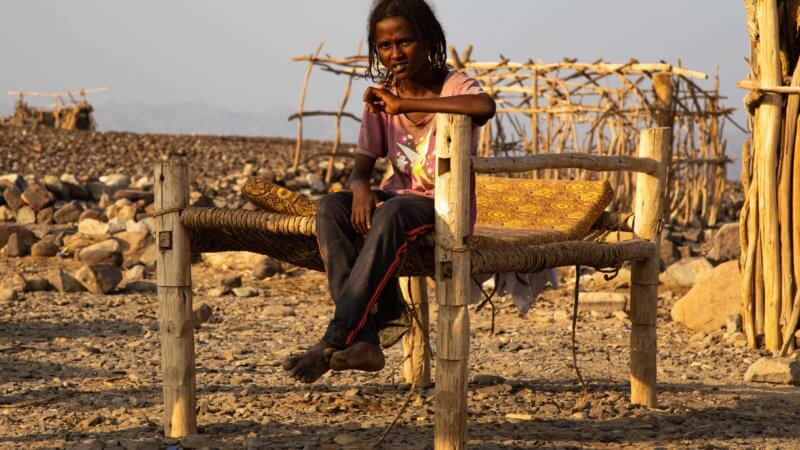 An Afar girl with dreadlocks sat on a wooden bed in the open air.