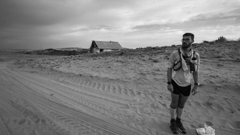 Black and white photo of Jamie Maddison stopping for a rest break near to an abandoned farmstead in Kazakhstan.