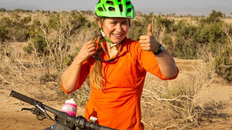 Mountain biker with orange top and green helmet gives a thumbs-up.