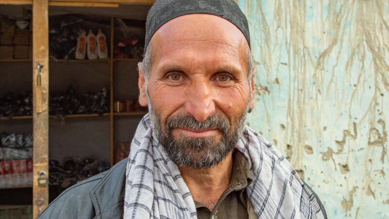 Friendly-looking shopkeeper with a cap on his head and loose white scarf around neck.