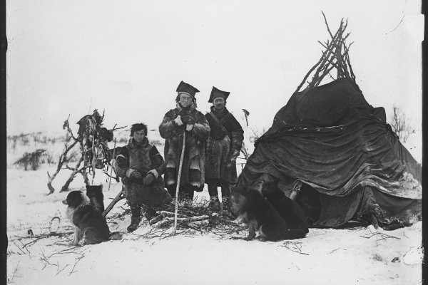Historical photo of three Sami and their dogs stood next to their lavvu traditional wooden tent.